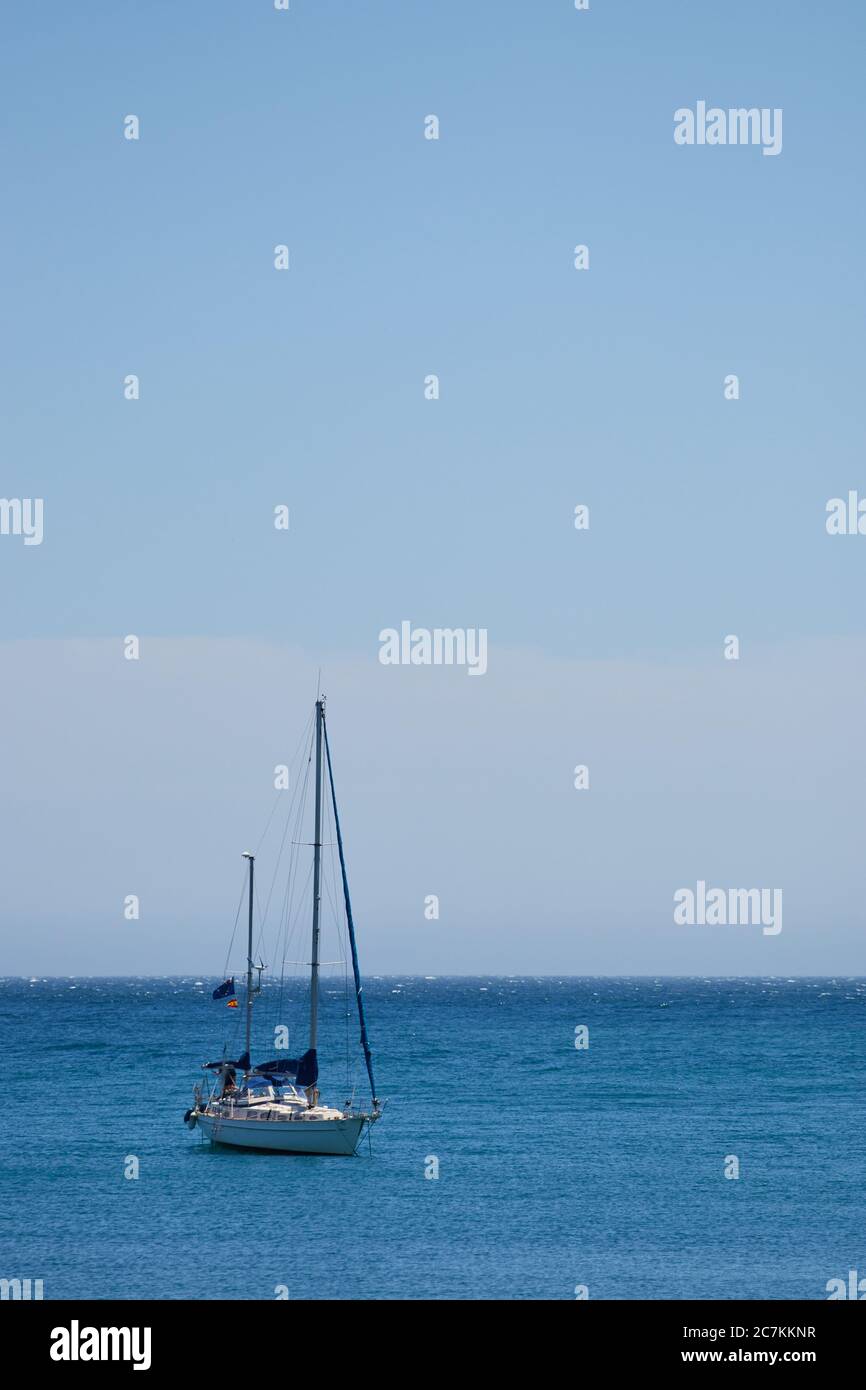 Vertikale Aufnahme mit einem Segelboot links vom Rahmen auf einem ruhigen Meer mit einem hellblauen wolkenlosen Himmel in malaga Stockfoto