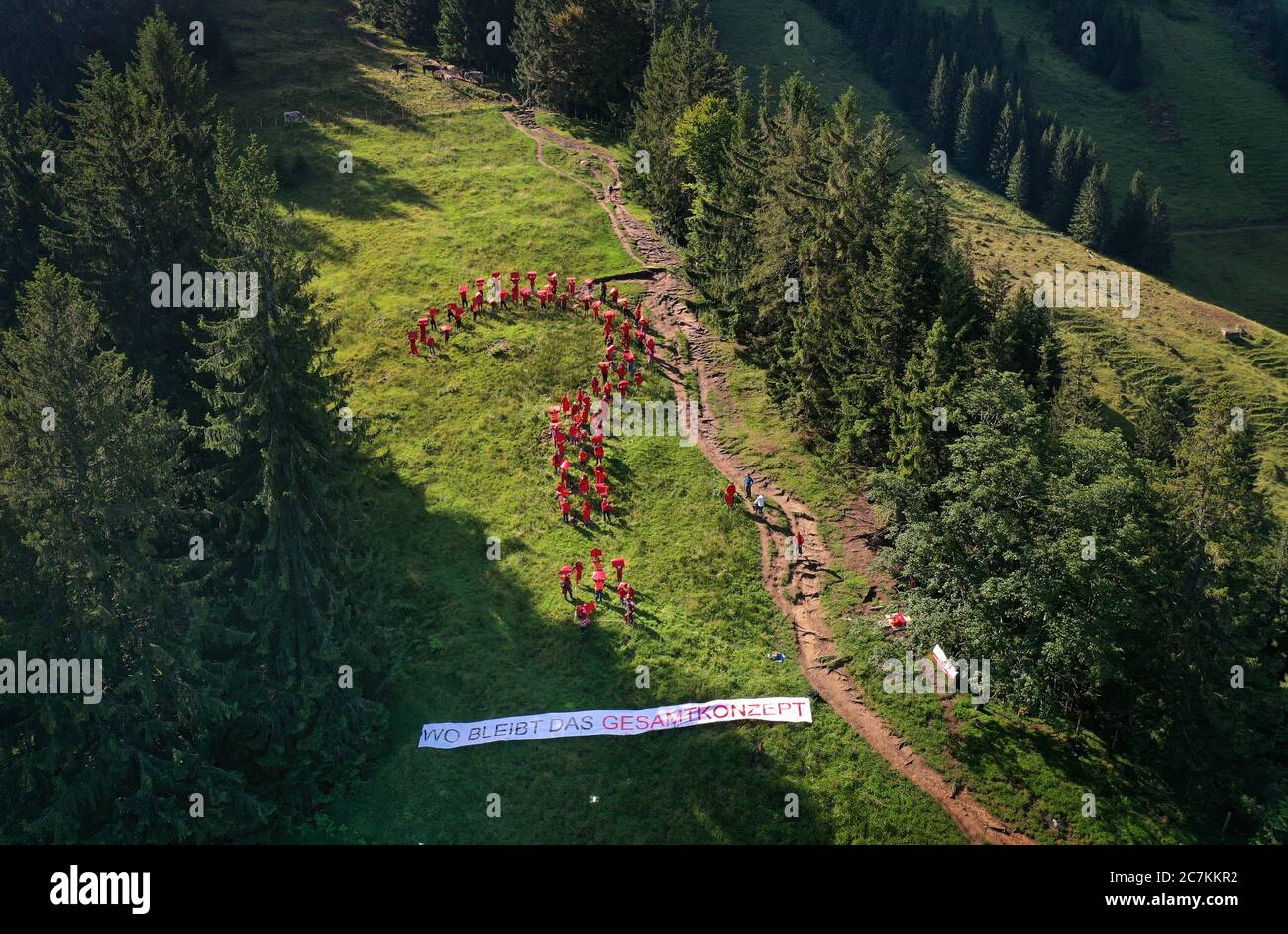 18. Juli 2020, Bayern, Rettenberg: Rund 80 Menschen in rot gekleidet stehen auf einer Bergwiese auf den Grünten in Form eines Fragezeichens über einem Plakat mit der Aufschrift "wo ist das Gesamtkonzept" (Luftaufnahme mit Drohne). Mit dieser Aktion demonstriert die Bürgerinitiative "Rettet den Grünen" gegen die Bergwelt-Pläne auf den Grünten. Die Veranstalter wollen darauf aufmerksam machen, dass aus ihrer Sicht ein festes Gesamtkonzept für die Bergwelt fehlt, das dort von einer Unternehmerfamilie geplant wurde. Foto: Karl-Josef Hildenbrand/dpa Stockfoto