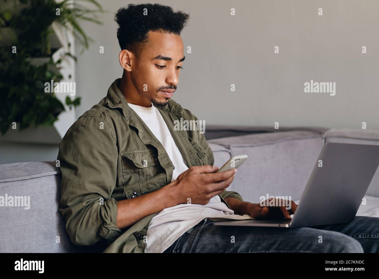 Junge ernsthafte afroamerikanische Kerl mit Laptop nachdenklich mit Handy in Co-Working-Raum Stockfoto