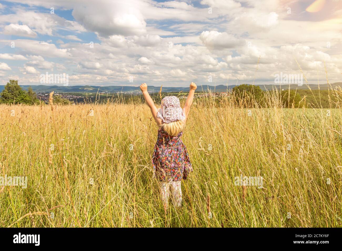 Nettes kleines blondes Mädchen, das im Sommer auf einem Feld steht und ihre Arme voller Freude hebt Stockfoto