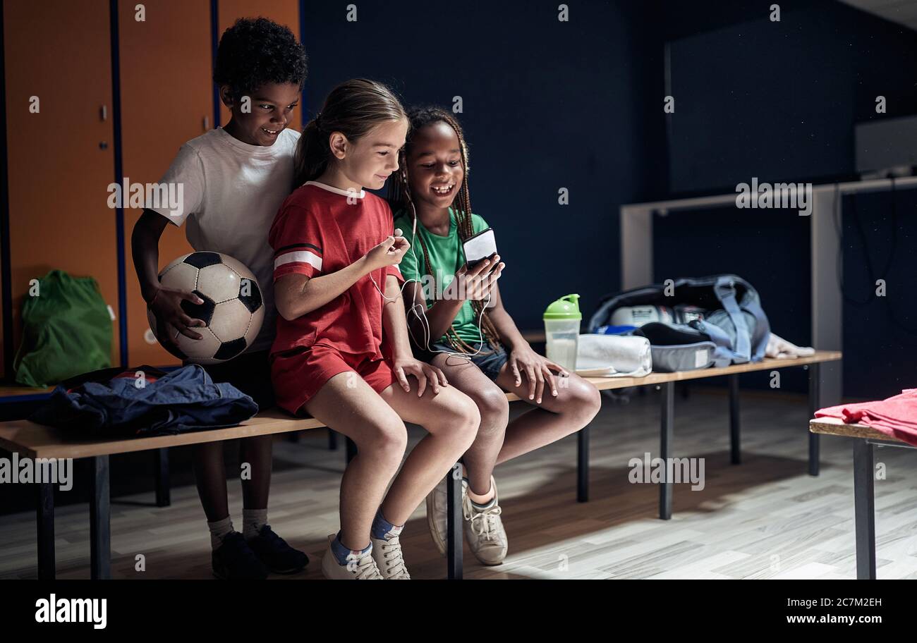 Lächelnde Kinder sitzen in einer Umkleidekabinen und hören Musik am Telefon vor dem Training. Stockfoto