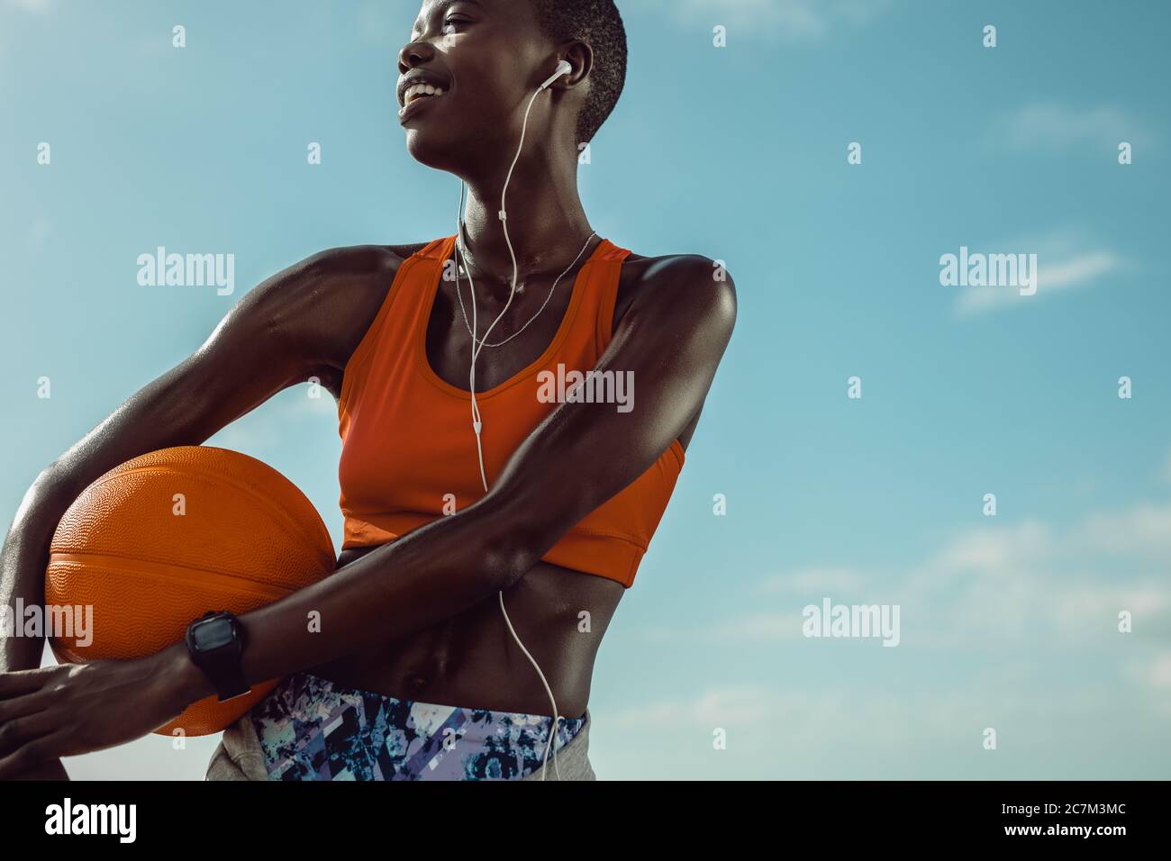 Sportlerin, die im Freien steht und einen Basketball hält. Frau mit einem Basketball, der wegschaut und gegen den Himmel lächelt. Stockfoto