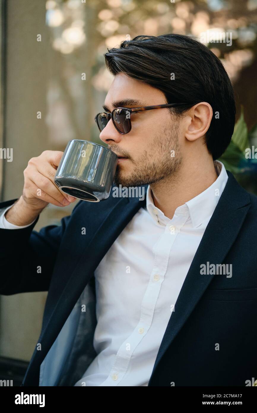 Junge attraktive bärtige Brünette Mann in weißem Hemd, klassische Jacke und Sonnenbrille verträumt neben beim Kaffee trinken auf der Straße der Stadt Stockfoto