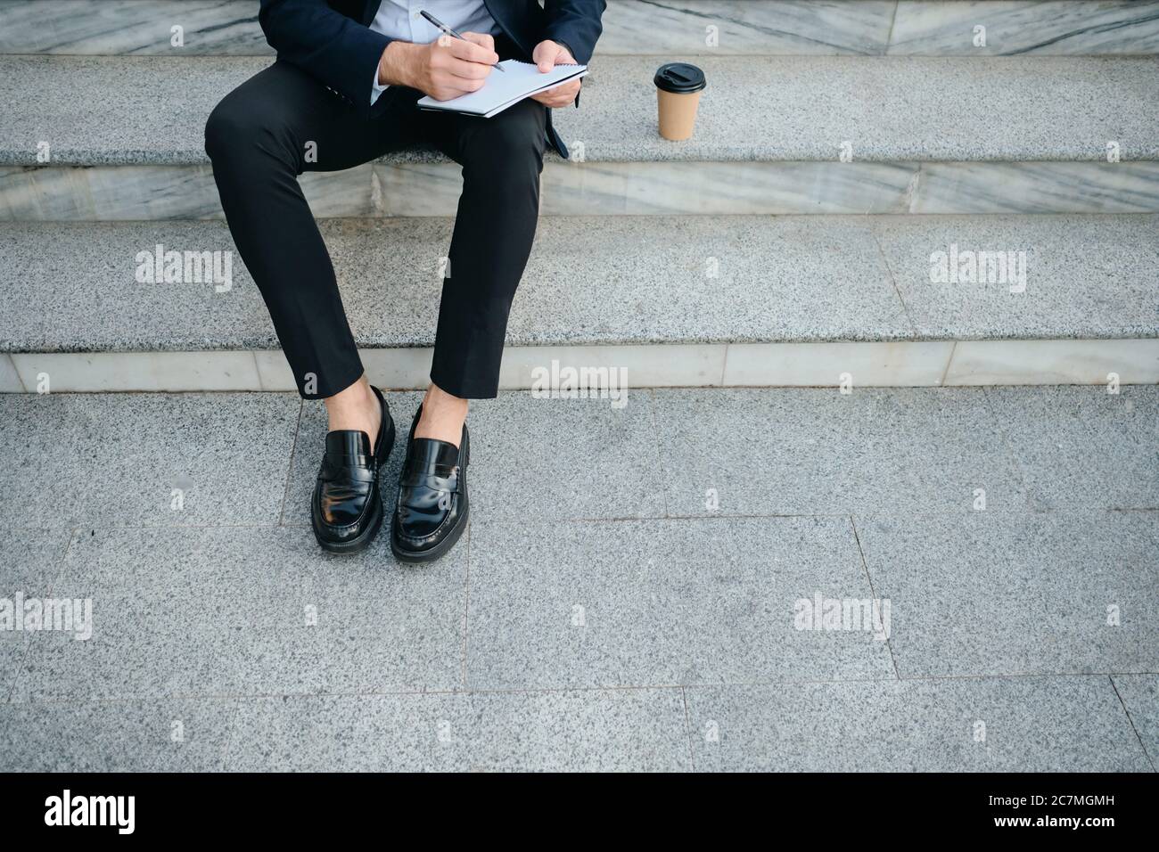 Nahaufnahme Mann im klassischen Anzug sitzt auf Treppen im Freien und Notizen in Notizblock mit einer Tasse Kaffee in der Nähe zu gehen Stockfoto