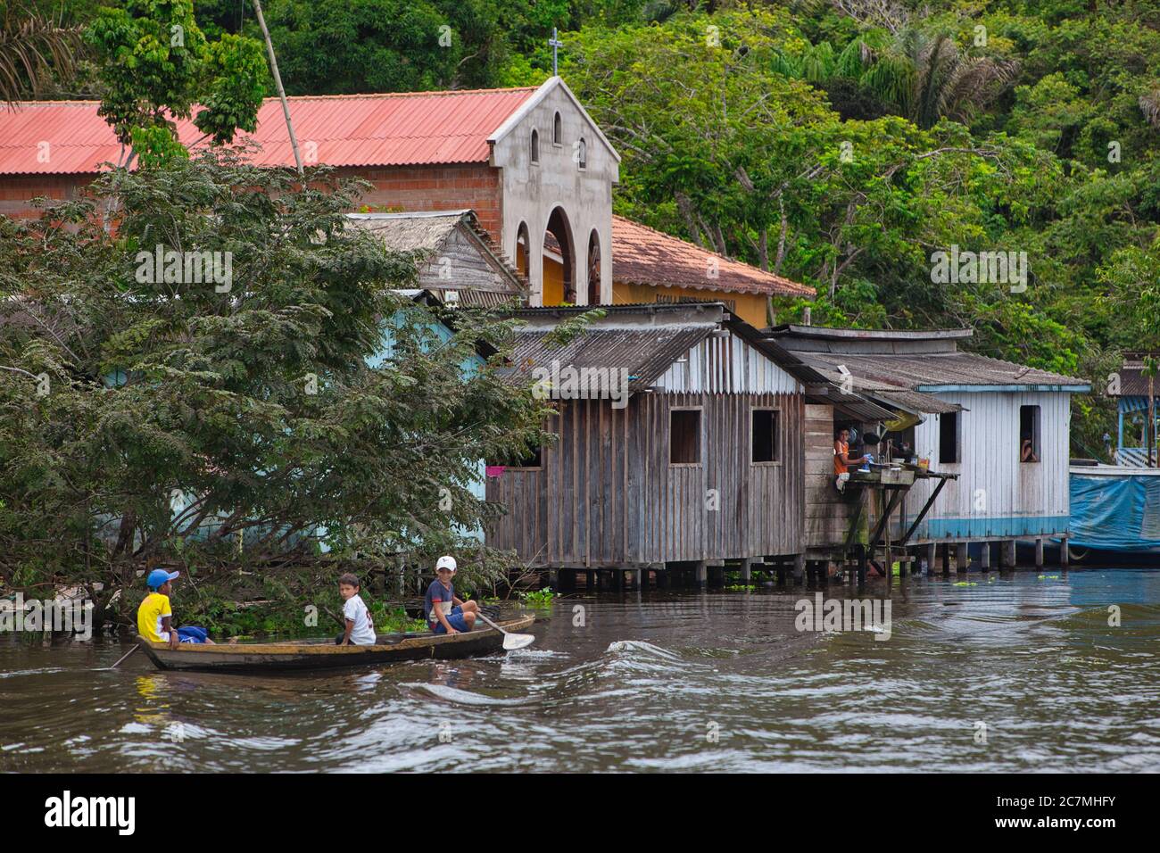 Drei Kinder paddeln ihr eigenes offenes Holzkanu mit Church Beyond im Dorf Boca De Valeria am Amazonas, Bundesstaat Amazonas, Brasilien Stockfoto