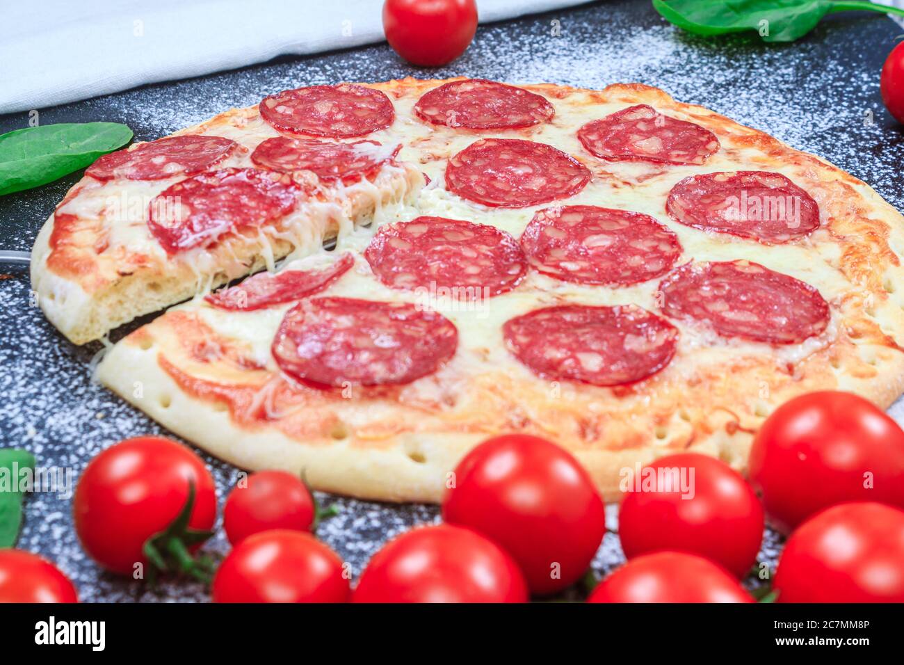 Pepperoni Pizza eine Scheibe abgeschnitten und Aufgang Käse Stretching goldenen Teig, Mehl reifen Kirschtomate Zweige mit Spinat auf einem schwarzen Tisch. Side vie Stockfoto