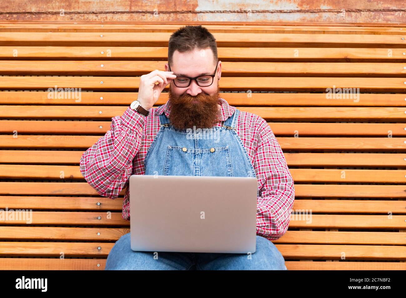 Nahaufnahme Porträt von angenehmen brutalen bärtigen Hipster Mann in blauen Overalls, kariertes Hemd arbeiten auf Laptop sitzen auf Bank im Freien. Distanzauftrag Stockfoto