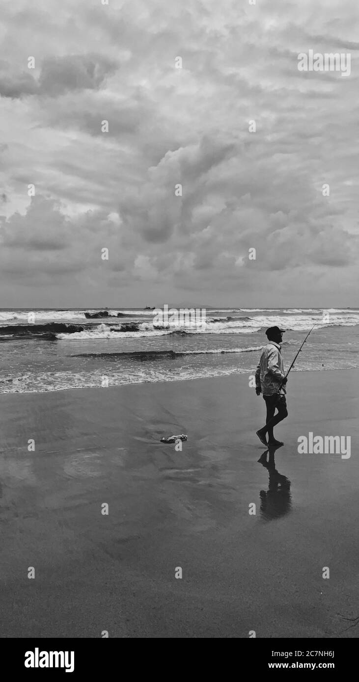 Vertikale Graustufenaufnahme eines Fischers, der entlang eines Sands geht Ufer Stockfoto