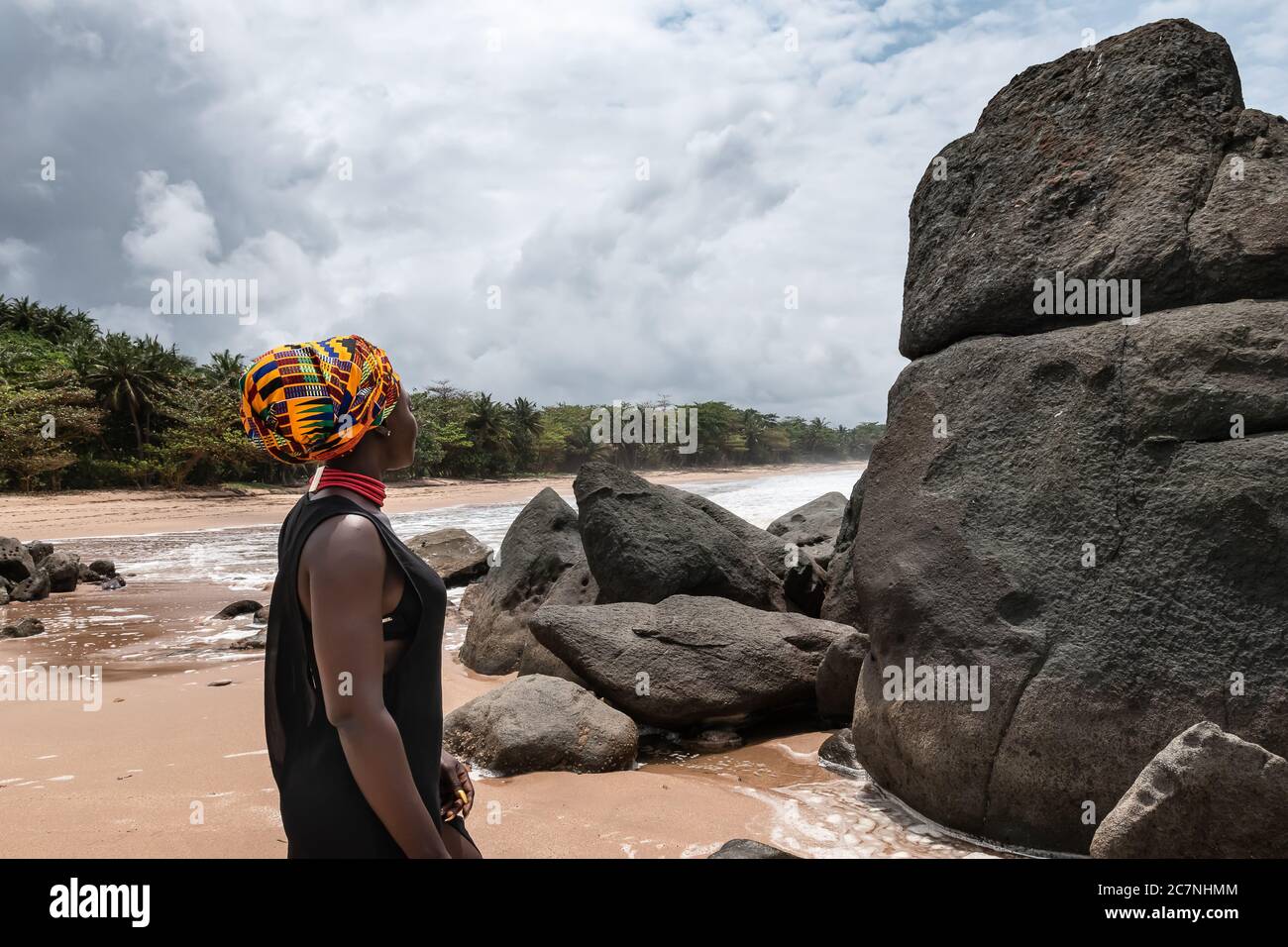 Afrikanische Frau steht an einem schönen Strand mit dunklen großen Steinen und sauberen Strand am Rande des Dschungels in Axim Ghana Westafrika Stockfoto