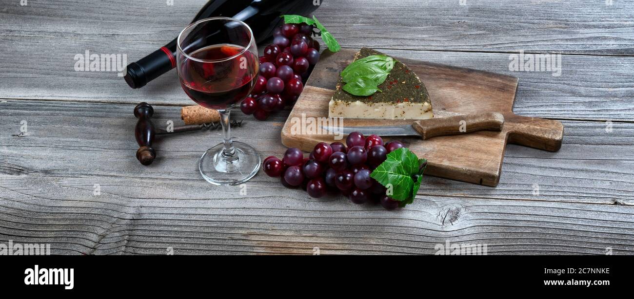 Glas Rotwein mit frischem Käsekeil plus Basilikumblätter und Trauben auf rustikalem Holztisch Stockfoto