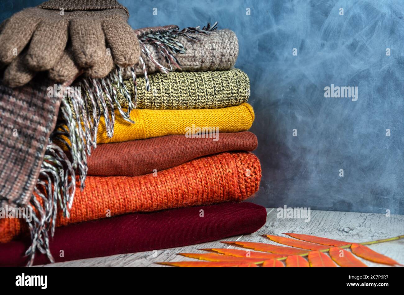 Stapel von gefalteten warmen Strickpullover für Damen in warmen Farben, karierten Schal, Strickhandschuhen und Herbstblättern auf grauem Hintergrund. Speicherplatz kopieren. Nahaufnahme Stockfoto