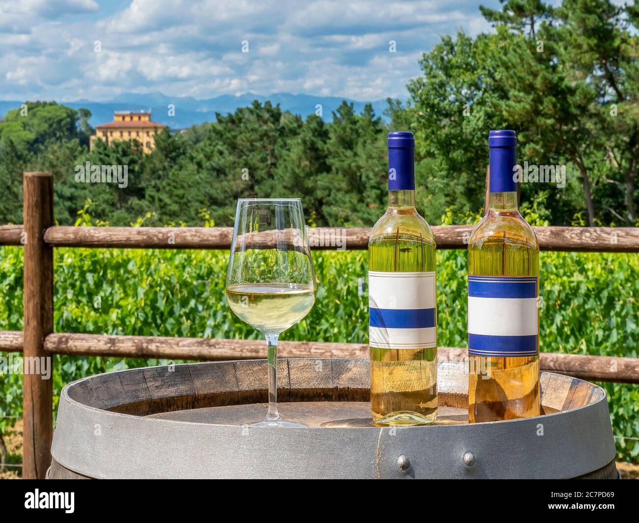 Zwei Flaschen und ein Glas Weißwein auf einem Holzfass mit der toskanischen Landschaft im Hintergrund, Italien, an einem sonnigen Tag Stockfoto