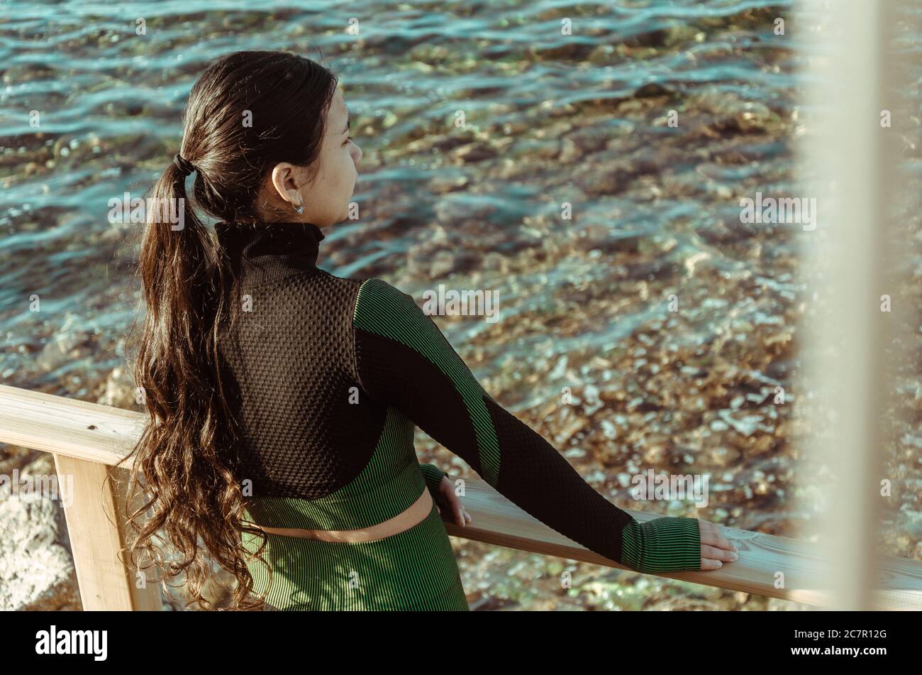 Junge hispanische Frau, die sich auf ein hölzernes Gesicht lehnt und schaut Am Meer unter dem Sonnenlicht Stockfoto