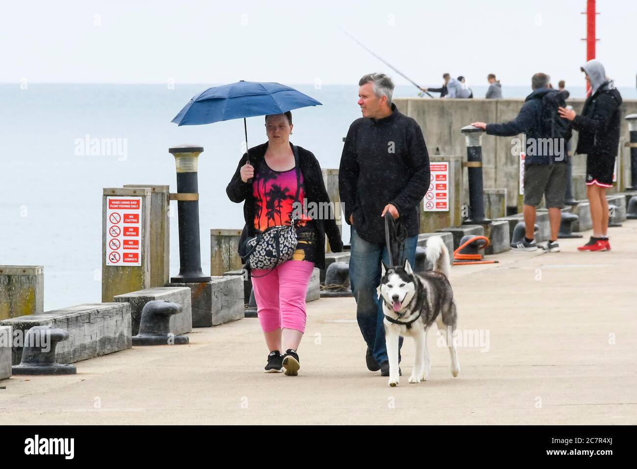 West Bay, Dorset, Großbritannien. Juli 2020. Wetter in Großbritannien. Hundespaziergänger am Pier mit einem Regenschirm im Badeort West Bay in Dorset an einem bewölkten Tag mit gelegentlichen leichten Regen. Bildquelle: Graham Hunt/Alamy Live News Stockfoto