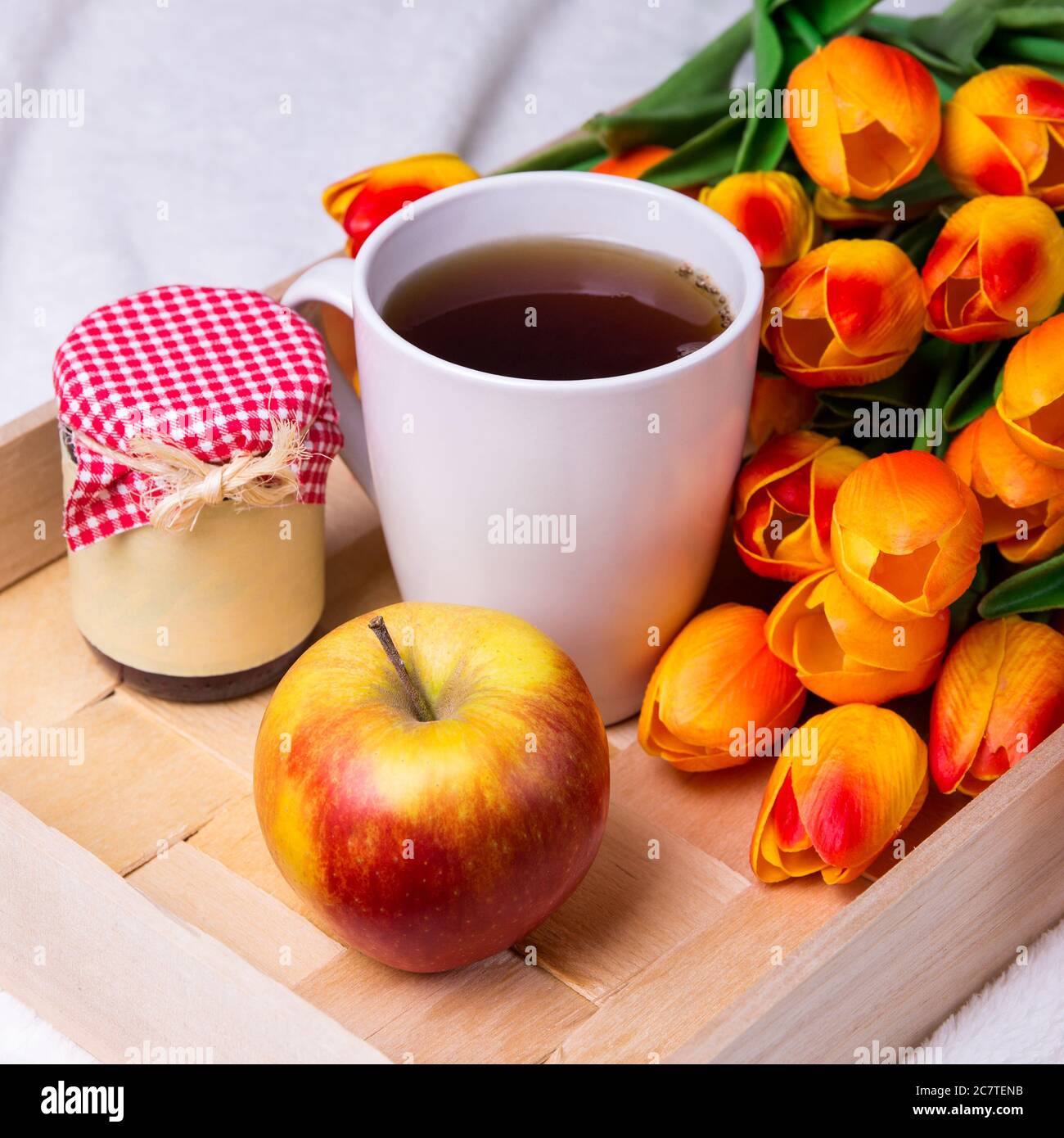 Nahaufnahme von Holztablett mit Tasse Tee, Glas Marmelade oder Honig, Apfel und Blumen Stockfoto