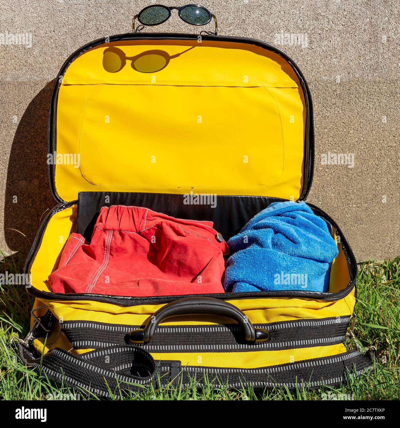 Ein gelber Koffer auf einer grünen Wiese ist mit einem Badeanzug und Strandtuch mit Sonnenbrille auf ihm gefüllt Stockfoto