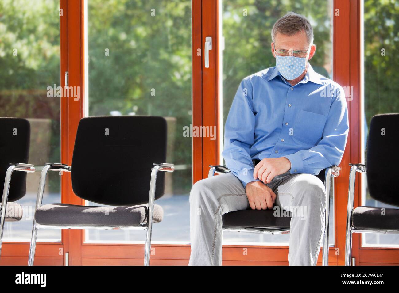 Trauriger oder müder reifer Mann mit Gesichtsmaske sitzt an einem sonnigen Tag in einem Wartezimmer eines Krankenhauses oder eines Büros Stockfoto