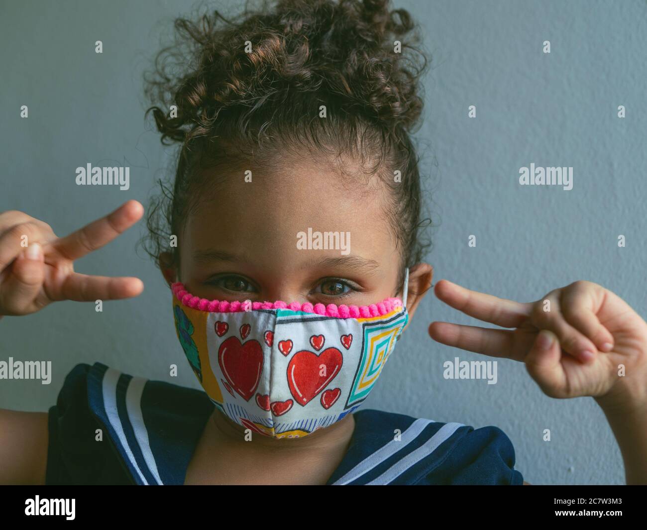 Ein kleines Mädchen mit einer Maske aus Herzen blickt mit den Fingern auf die Kamera mit dem Zeichen des Friedens Stockfoto