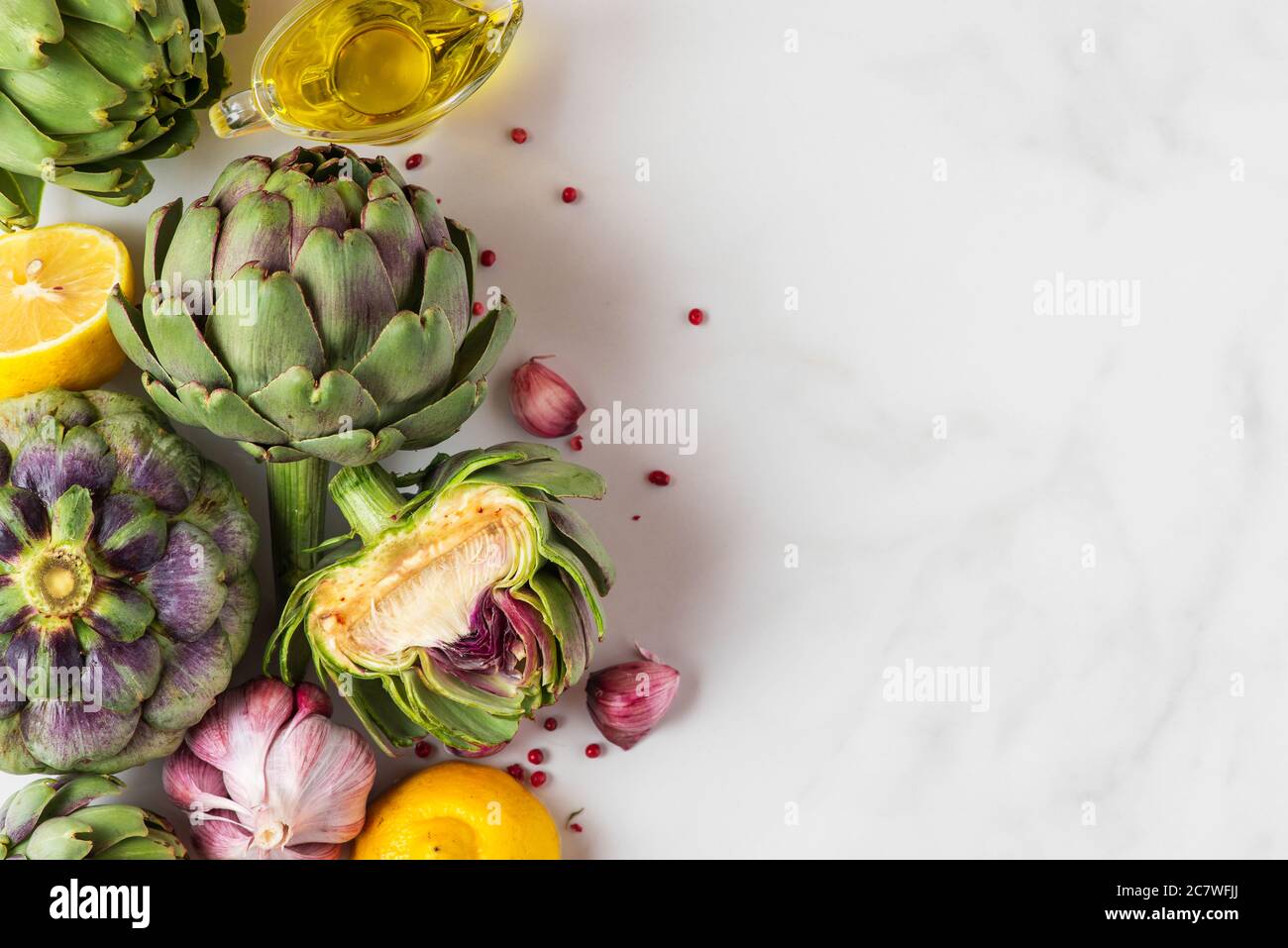 Frische reife Artischocken, Zitrone und Knoblauch mit Olivenöl und Pfeffer auf weißem Marmortisch. Gesundes veganes Kochkonzept. Flach liegend. Draufsicht mit c Stockfoto