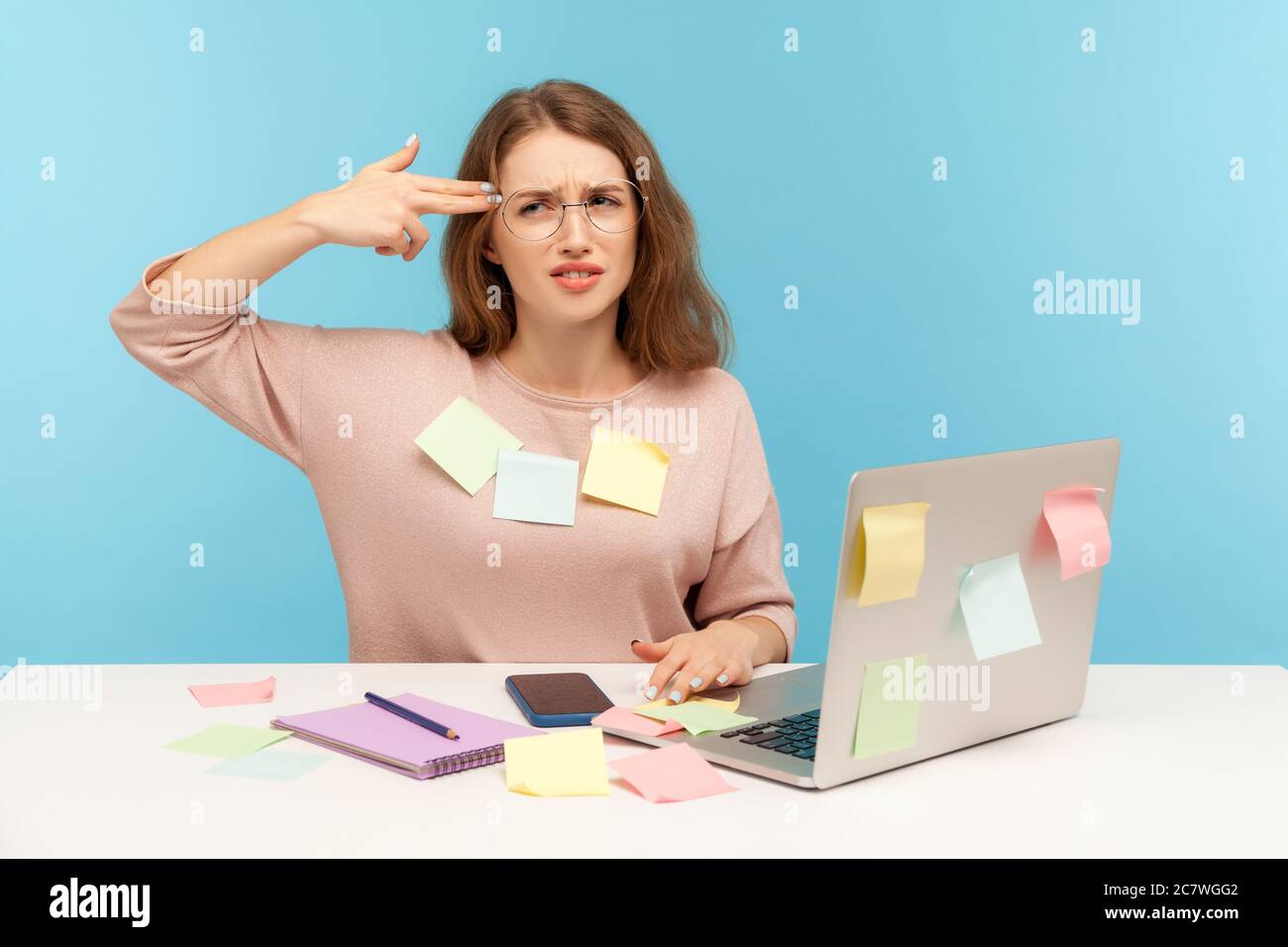 Deprimiert überarbeiteten Frau Angestellter in Nerd Brillen sitzen am Arbeitsplatz, alle mit Haftnotizen bedeckt, schießen sich mit Fingerpistole Selbstmord g Stockfoto