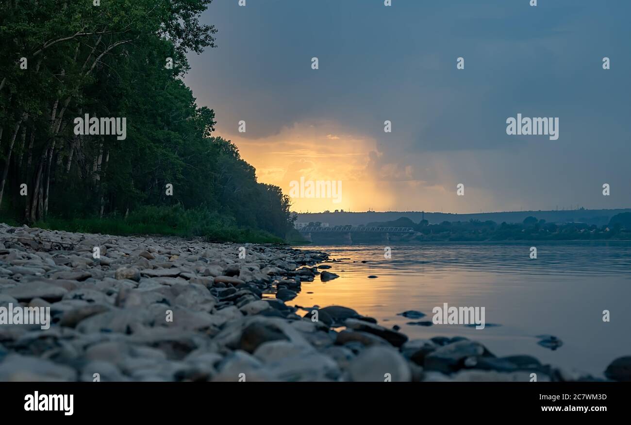 Abendlandschaft eines felsigen Ufer, ein Strand am Fluss vor dem Hintergrund eines bewölkten Regenhimmel mit einem schönen feurigen hellen sonnigen Sonnenuntergang Stockfoto