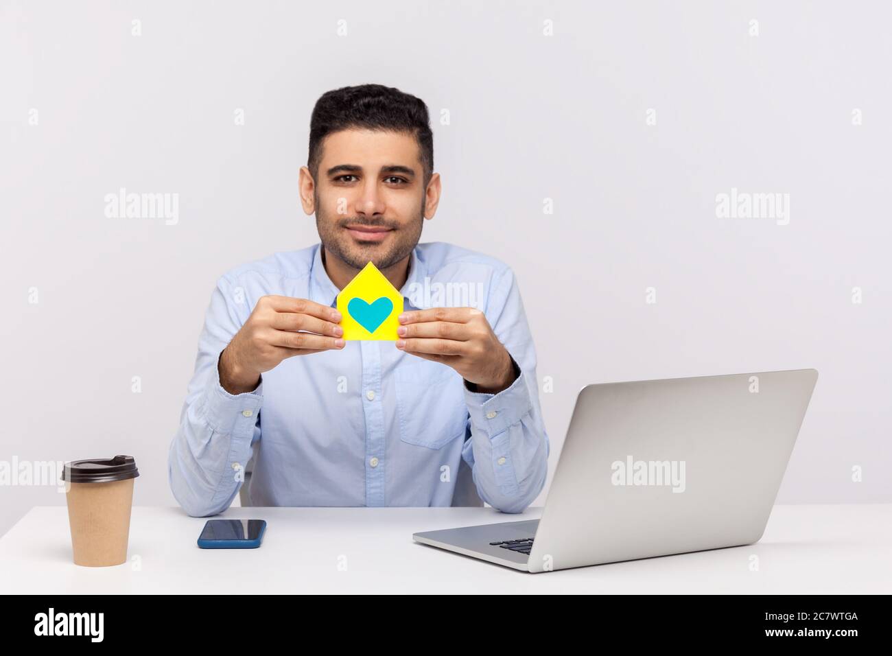 Portrait von glücklichen Immobilienmakler Mann sitzen in Büro-Arbeitsplatz und halten Papierhaus mit Herz im Inneren, lächelnd freundlich, Werbung von purc Stockfoto