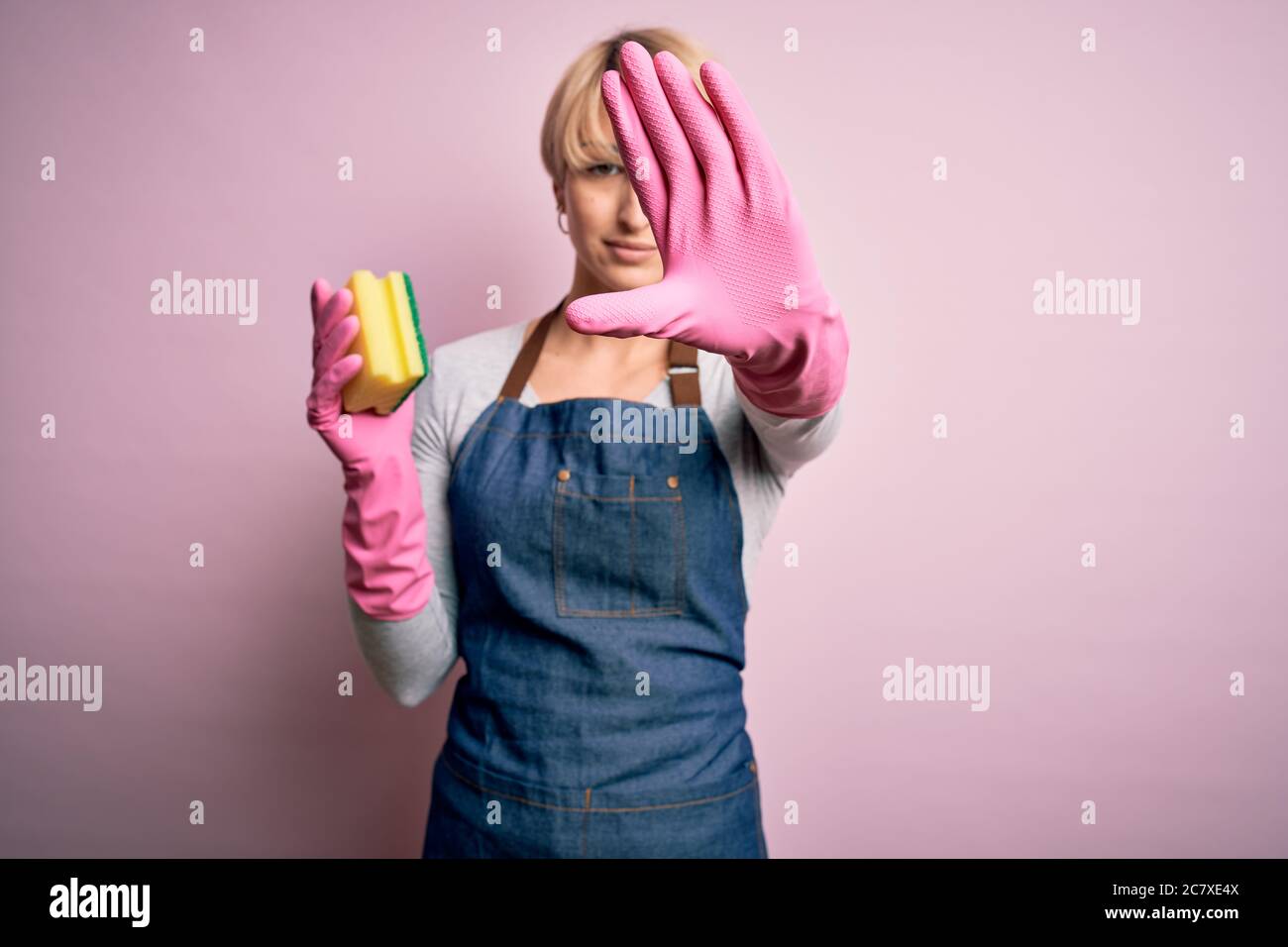 Junge blonde Putzfrau mit kurzen Haaren trägt Schürze und Handschuhe halten Peeling Geißel mit offener Hand tun Stop-Zeichen mit ernst und zuversichtlich ex Stockfoto