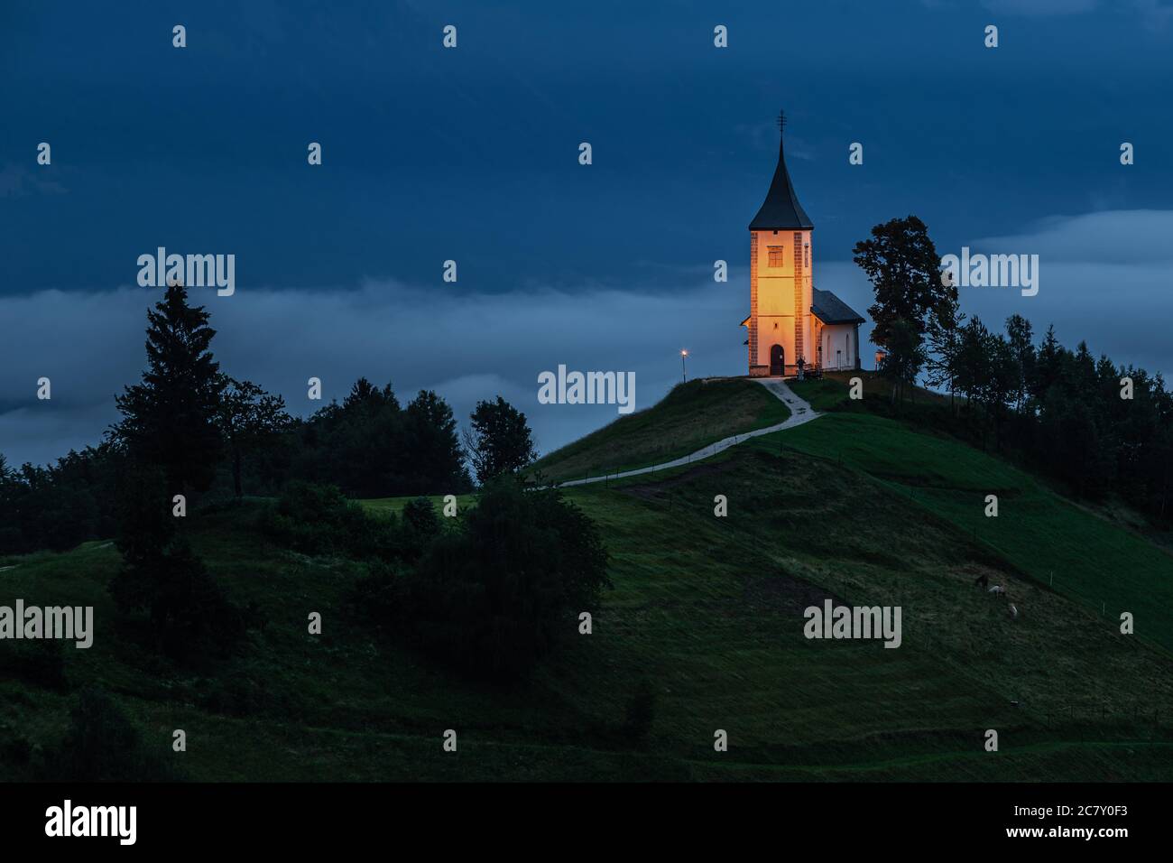Jamnik, Slowenien - Blaue Stunde in Jamnik mit beleuchteten St. Primoz Kirche in einem nebligen Morgengrauen. Die Julischen Alpen im Hintergrund Stockfoto