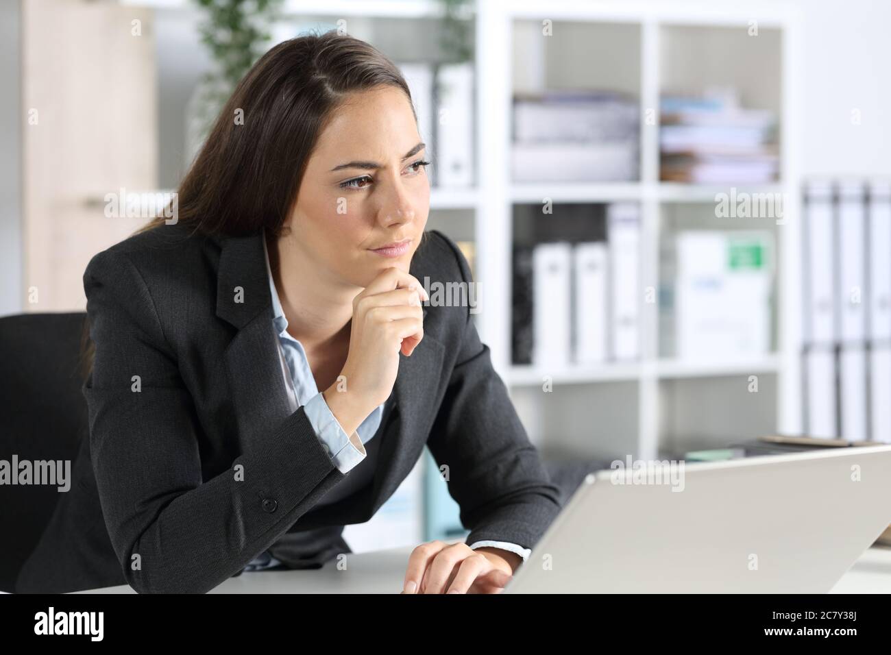 Nachdenkliche Frau, die mit einem Laptop im Büro auf ihrem Schreibtisch sitzt, denkt, sie schaue einfach nur aus Stockfoto