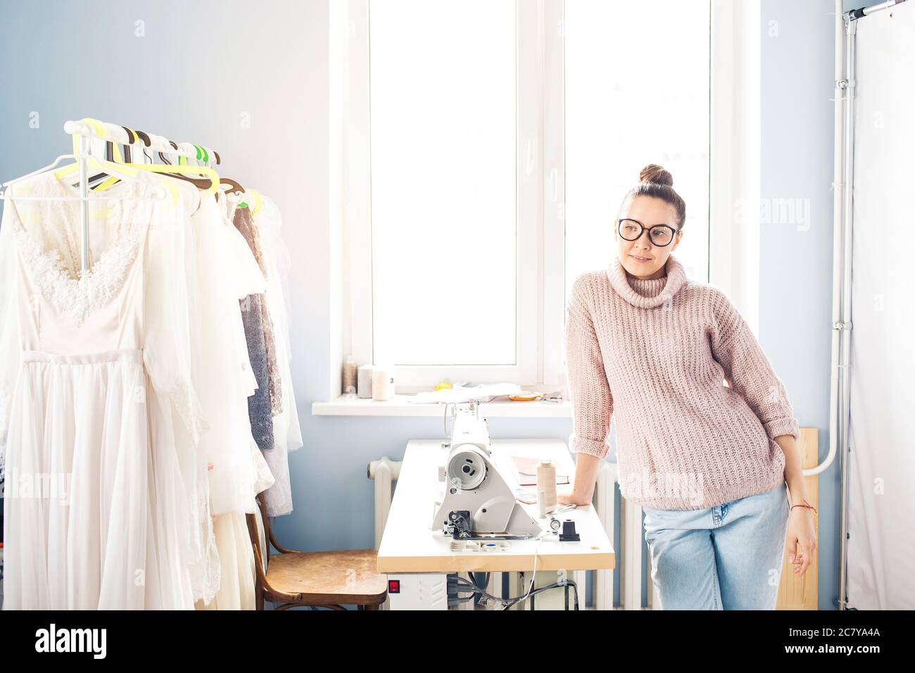 Schöne Designerin arbeitet im Schneiderstudio, schaut auf Kamera und lächelt, im Hintergrund ihr Kleid Stockfoto