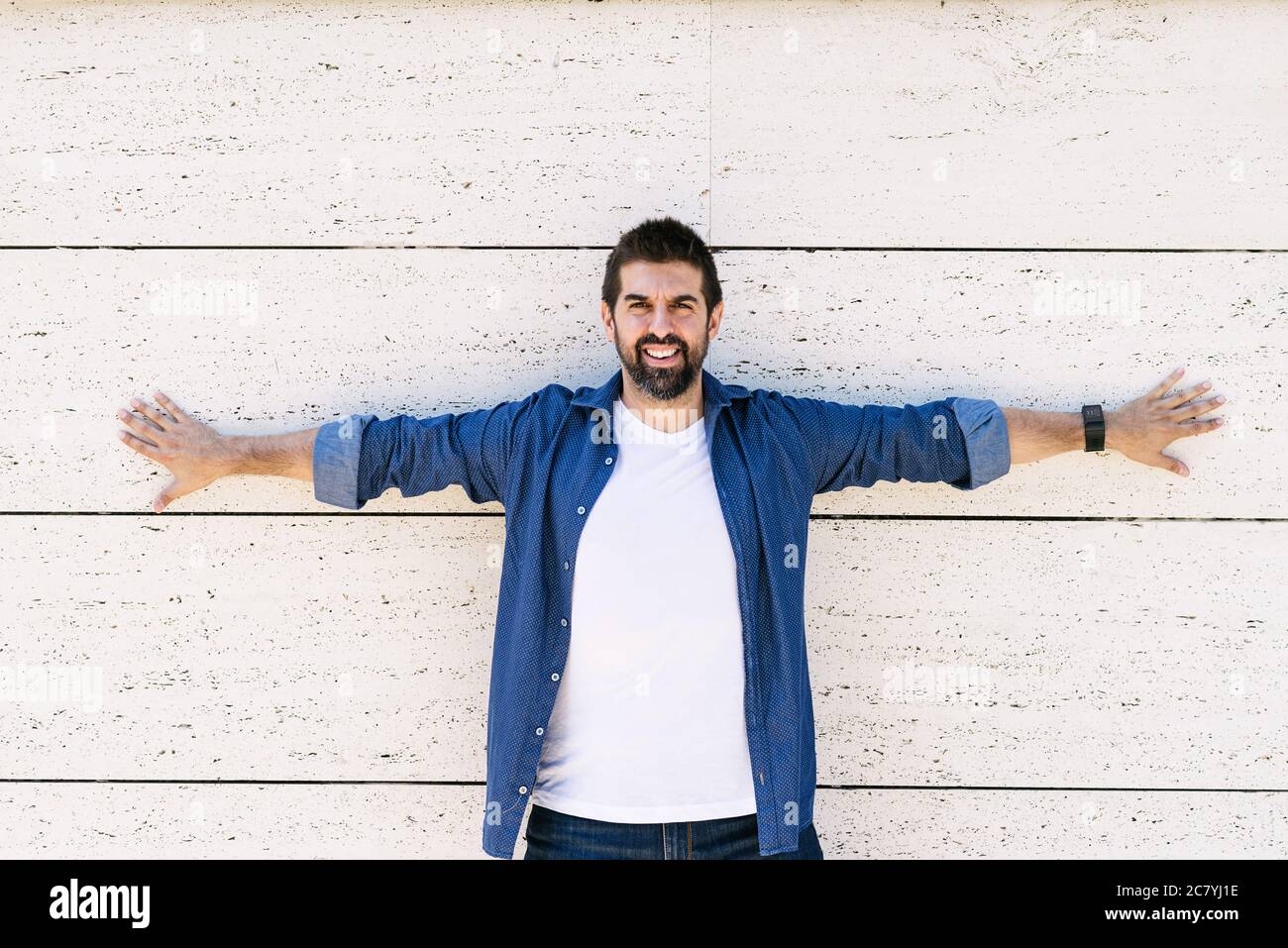 Bärtiger Mann, der mit gestreckten Armen an der Wand gelehnt ist und mit einem Lächeln auf die Kamera schaut Stockfoto