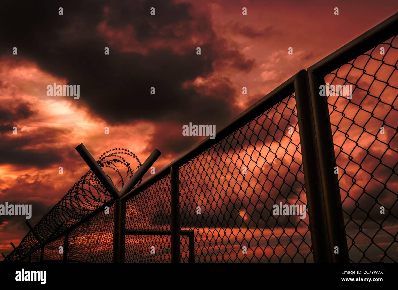 Sicherheitszaun der Militärzone oder privaten Bereich Zaun mit rotem Himmel und dunklen Wolken. Stacheldraht-Sicherheitszaun mit stürmischem Himmel. Barriere Grenze. Stockfoto