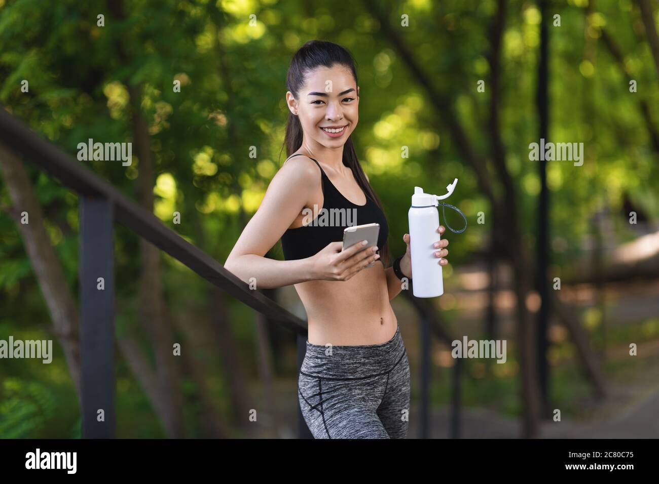 Lächelndes asiatisches Mädchen, das während des Trainings im Freien ruht, Smartphone und Trinkwasser benutzt Stockfoto