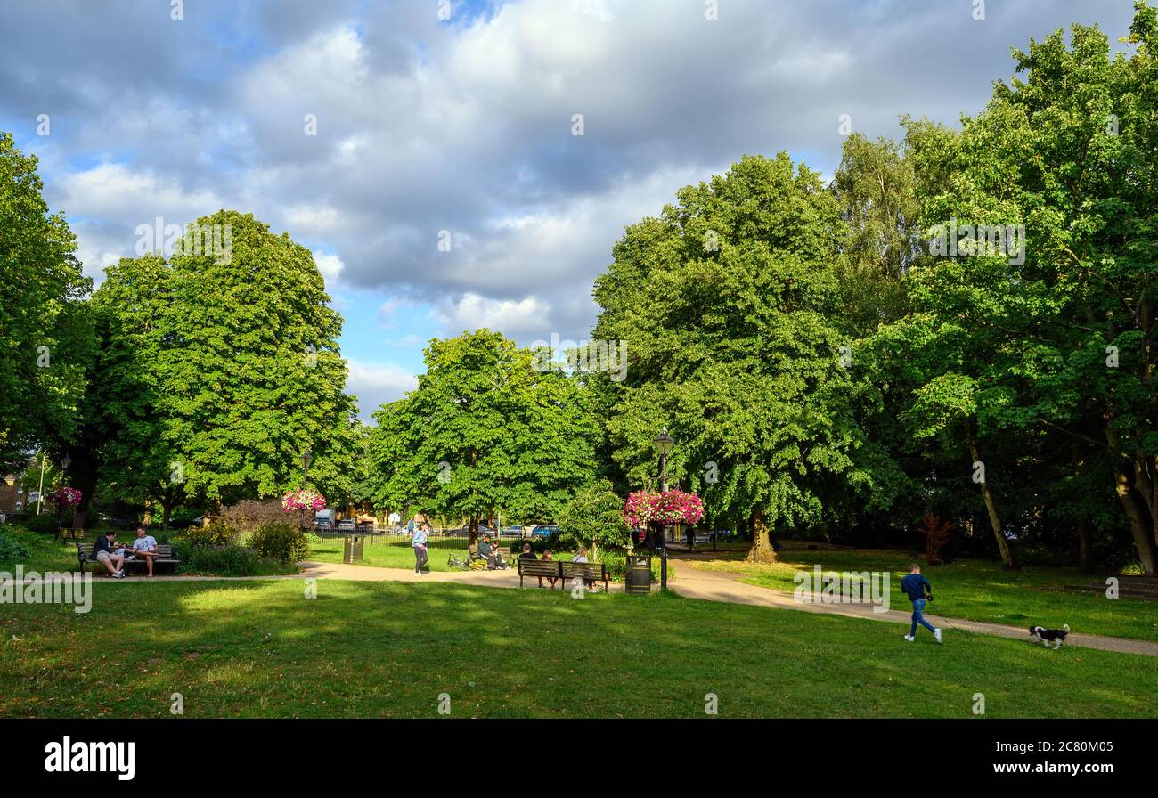 Beckenham (Großraum London), Kent, Großbritannien. Beckenham Grün mit Bäumen, Blumen und Gras. Die Leute sitzen auf Bänken und ein Junge läuft mit einem Hund. Stockfoto
