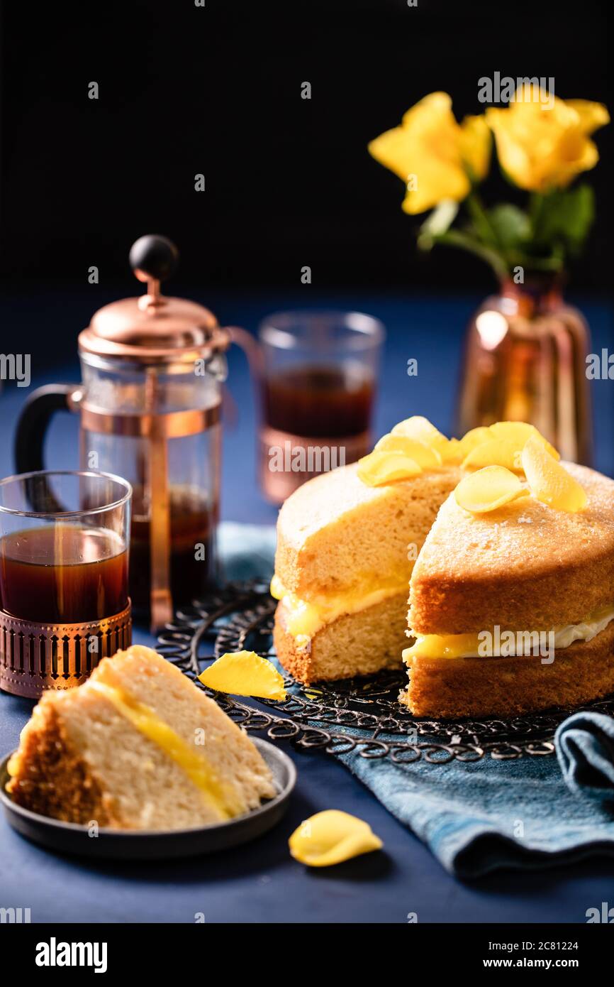 Dekadente Zitrone Biskuit mit gelben Rosenblättern mit frischem Kaffee auf tiefblauem Hintergrund serviert dekoriert Stockfoto