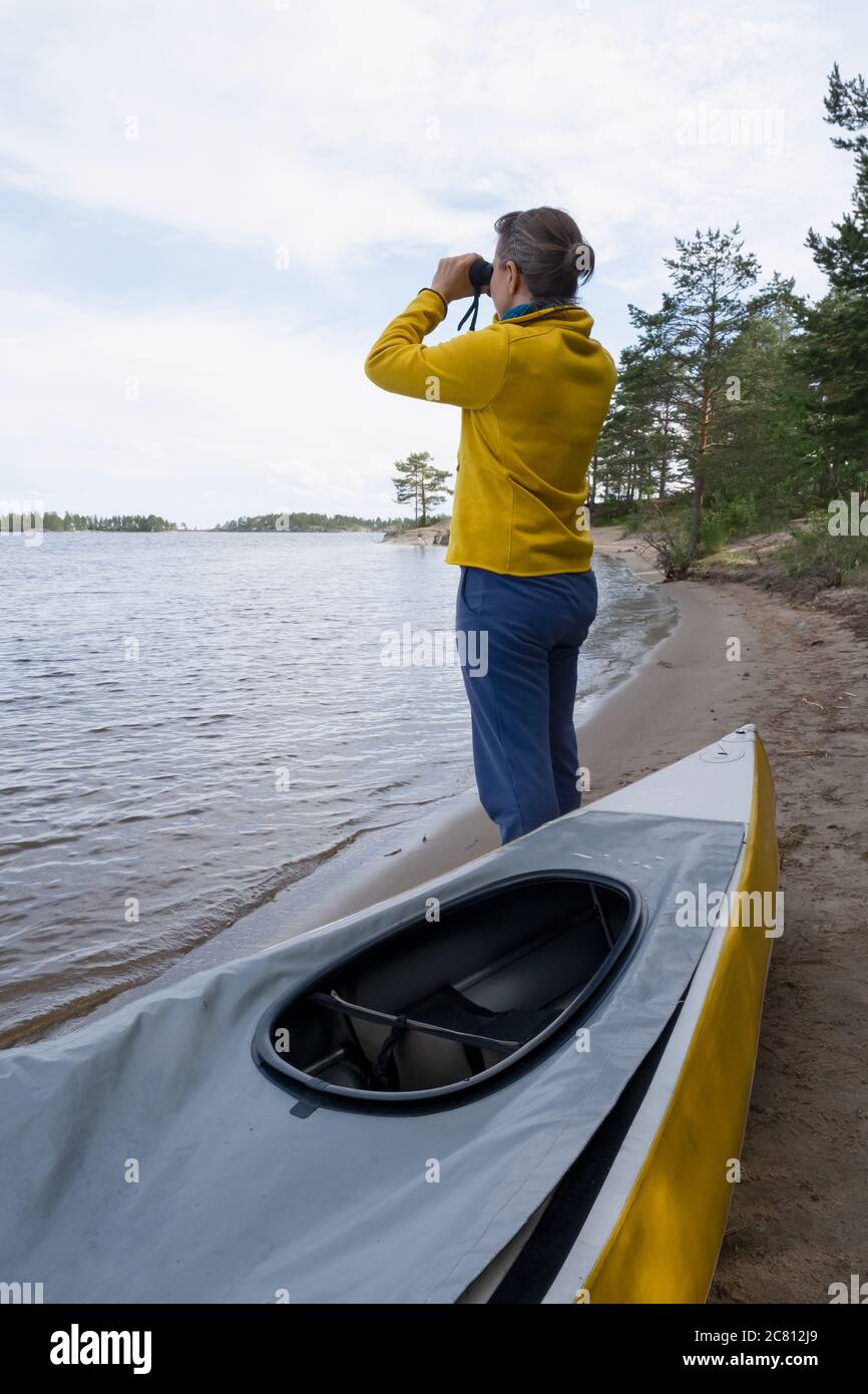 Frau, die in Fernglas weit entfernt in der Nähe von Kajakund Wasser des Sees steht. Stockfoto