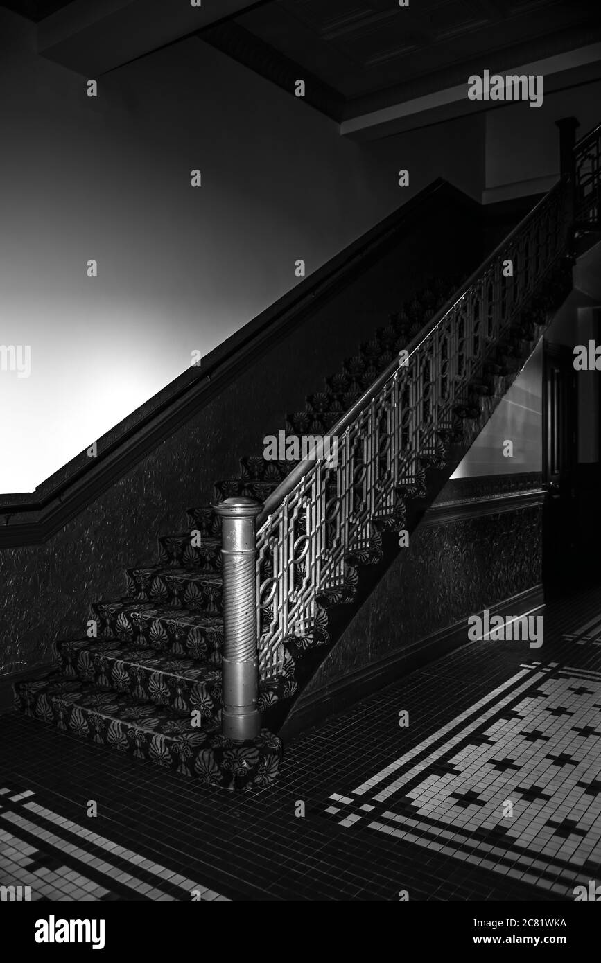 Im Mittelpunkt steht ein alter dunkler Handlauf einer Treppe mit geprägter Zinnvertäfelung, Pub-Teppich und Mosaikfliesen in dunkler Atmosphäre, in Schwarz und Weiß Stockfoto