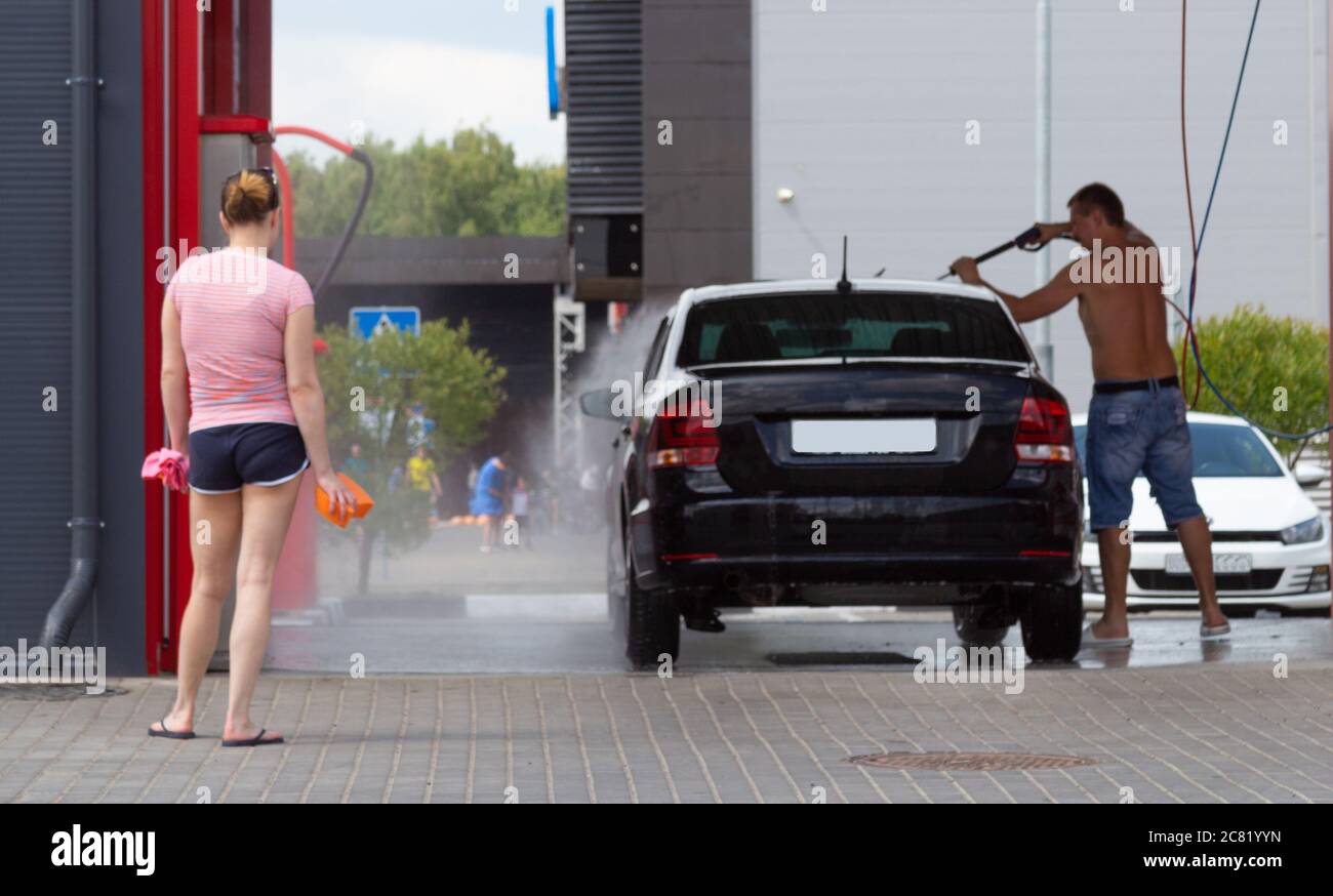 Ein Mann wäscht sein Auto mit einem Schlauch unter dem Druck von Wasser. Die Frau steht in der Nähe. Autowäsche Stockfoto