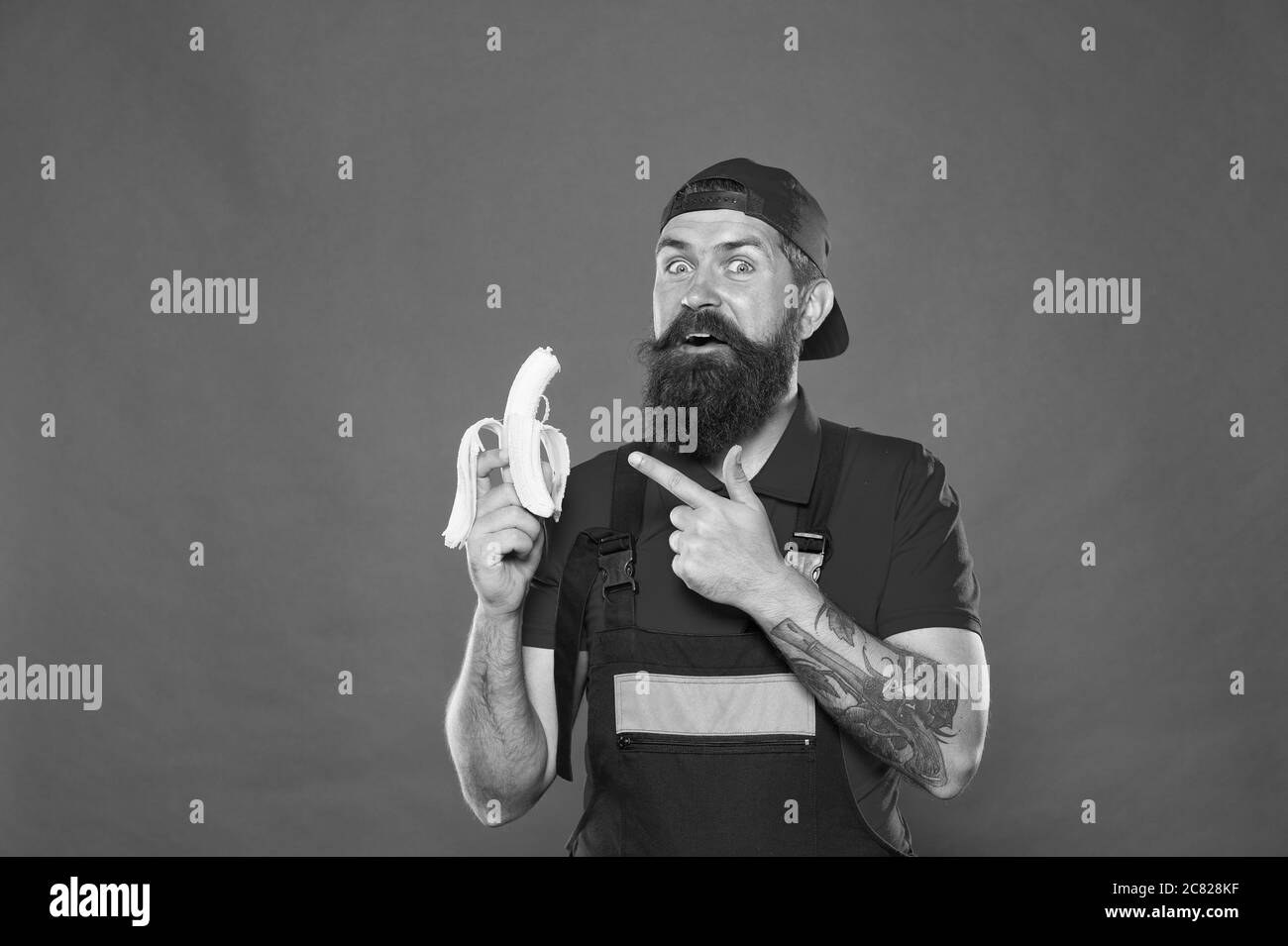 Hungriger Hipster. Lieblingssnack. Food Court. Mittagszeit. Bärtiger Mann in Uniform und Mütze essen Banane. Arbeiter Baumeister Mechaniker essen Banane. Frisches Obst. Ein gesundes Leben. Leckeres Essen. Biss Banane. Stockfoto