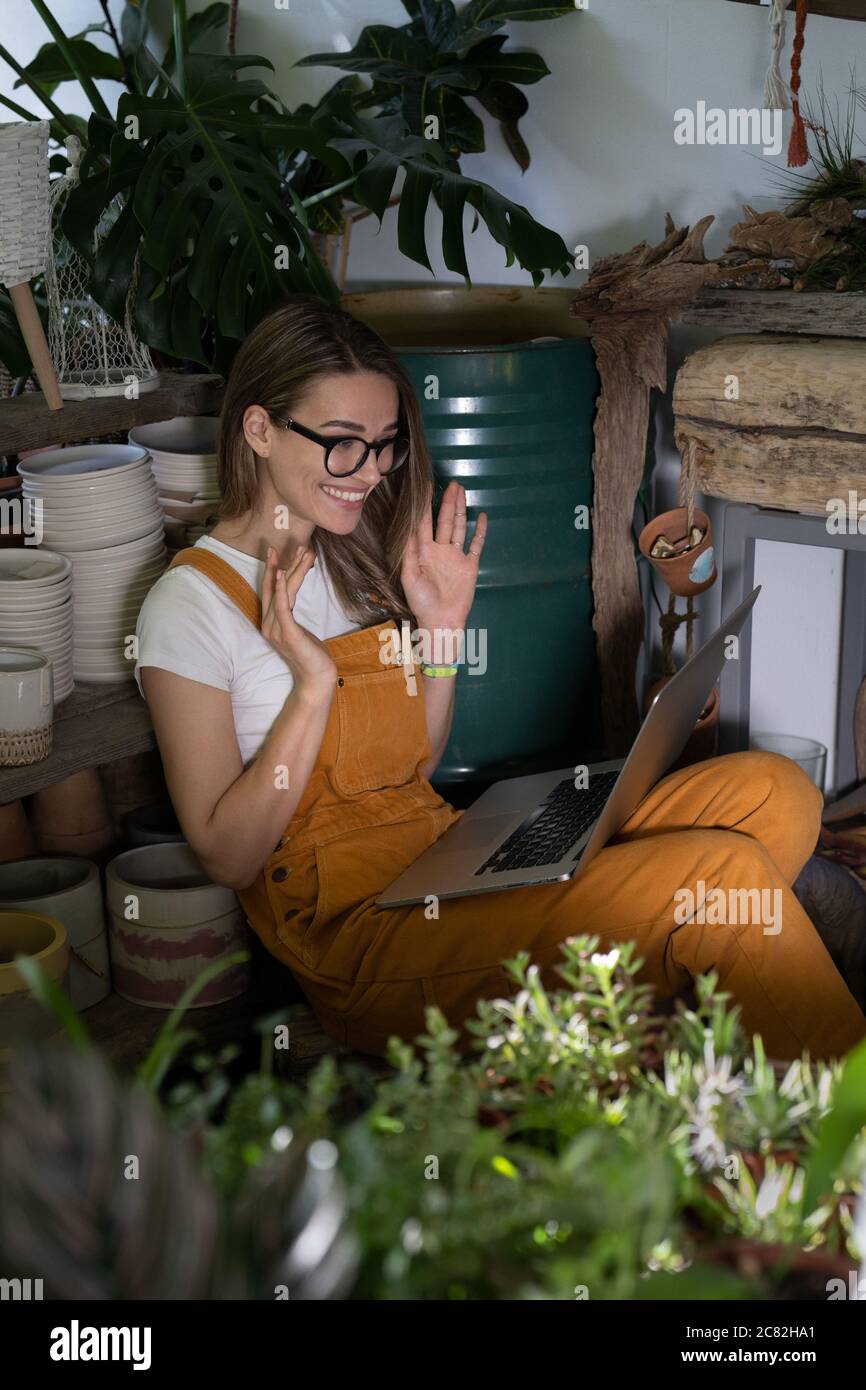 Nette Frau Floristin in Brille tragen orange Overalls, sitzen auf dem Boden in Blumenladen, ruhen, mit Laptop nach der Arbeit, lächeln und sprechen auf vid Stockfoto
