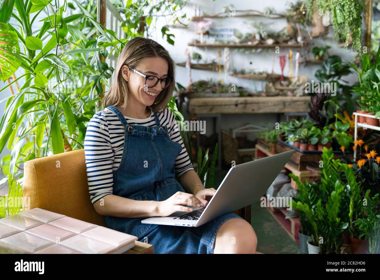 Frau Gärtnerin in Brille tragen blaue Jeans Overalls, sitzen auf Stuhl im Gewächshaus, mit Laptop nach der Arbeit, kommuniziert im Internet mit Kunden su Stockfoto