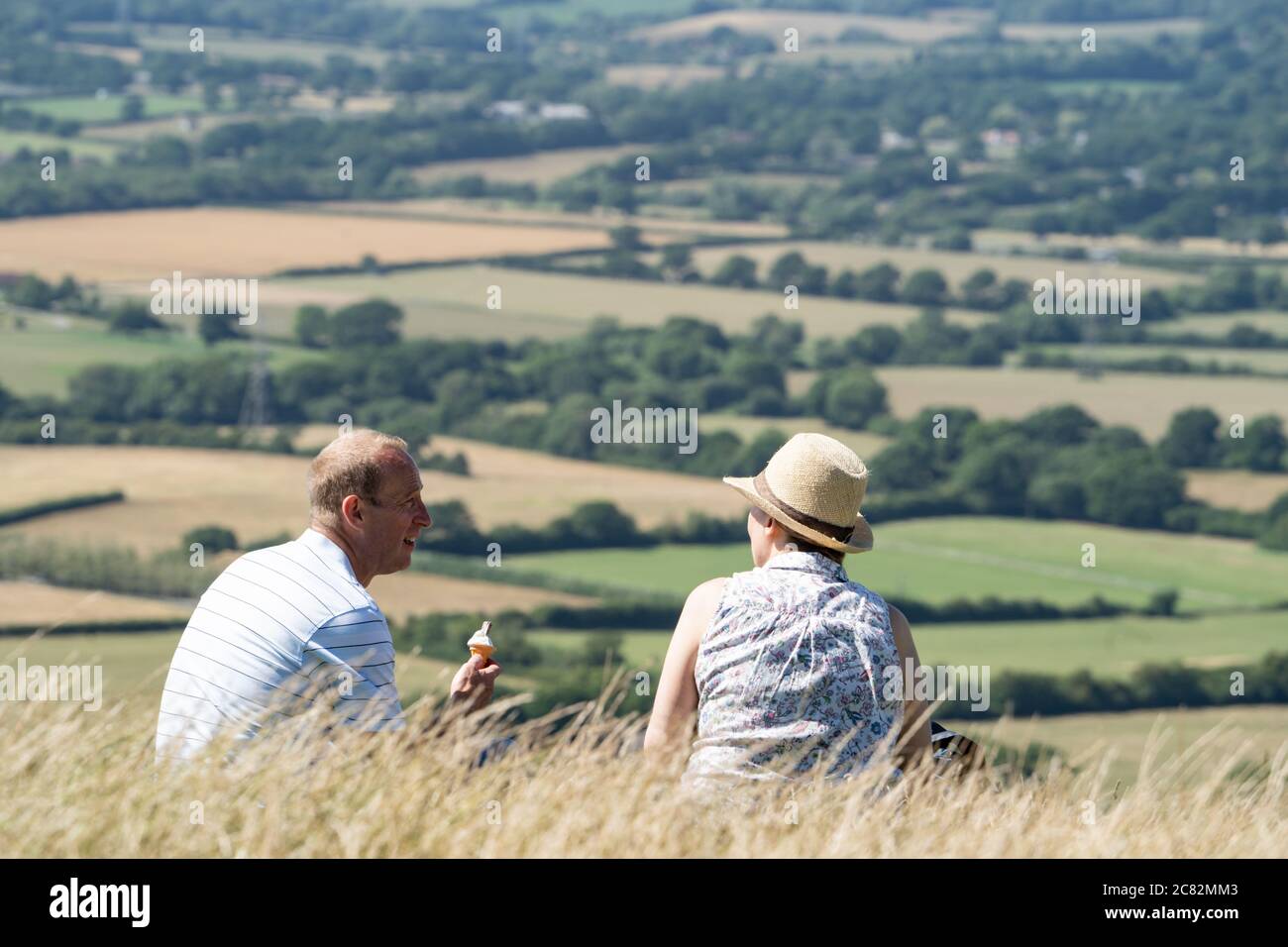 Ehepaar mittleren Alters sitzt auf einem Hügel am Devil's Dike Valley, South Downs, Sussex, Großbritannien, essen Eis und reden Stockfoto