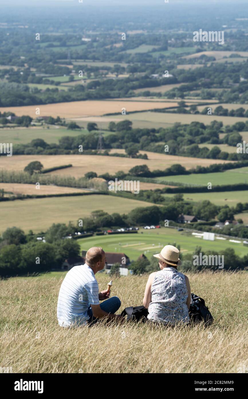 Ein Paar mittleren Alters, das auf einem Hügel im Devil's Dike Valley, South Downs, Sussex, Großbritannien, sitzt und Eis isst, überblickt den Preston Nomads Cricket Club, Fulking Stockfoto
