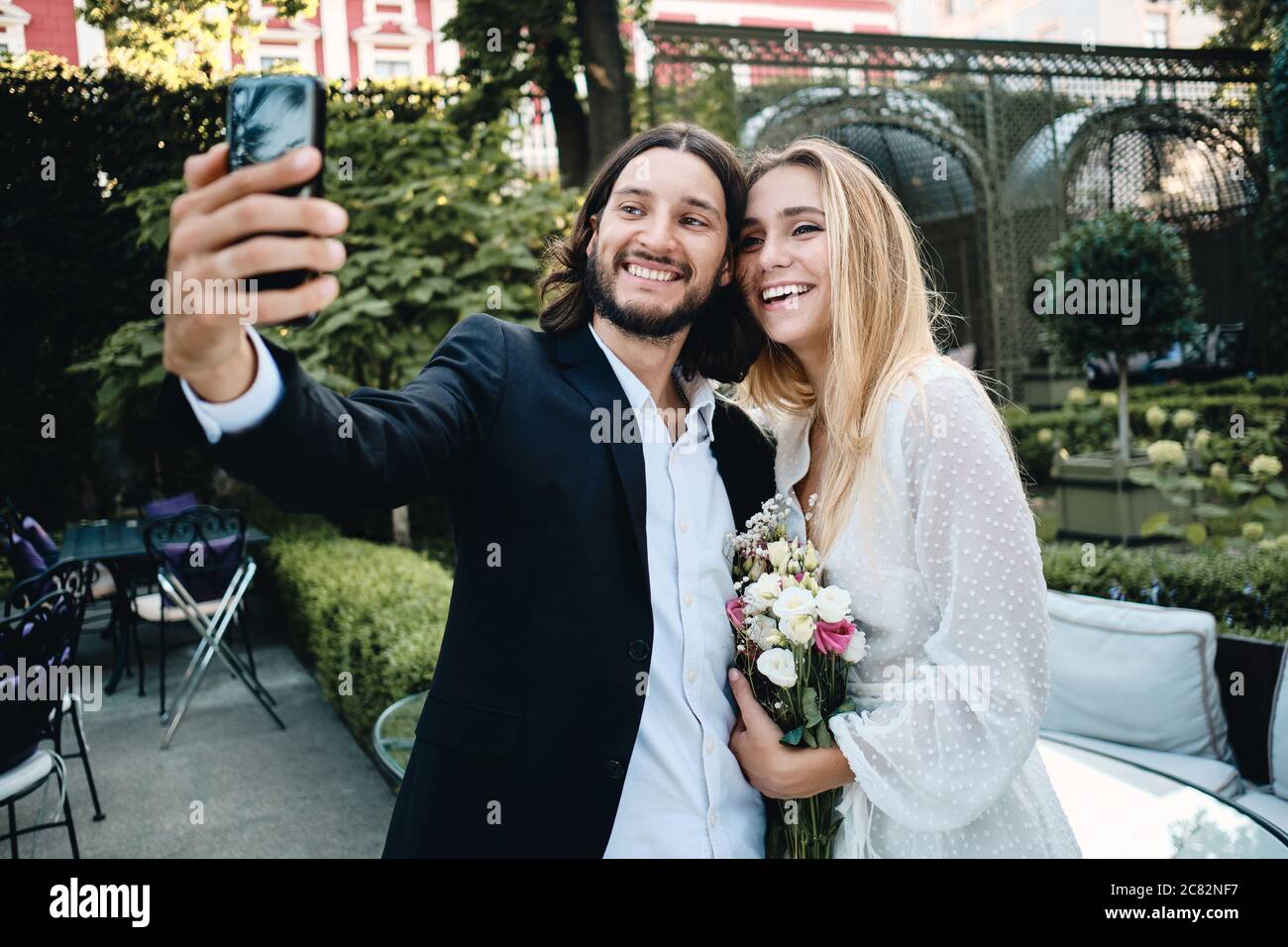 Junge fröhliche Bräutigam und Braut glücklich Selfie auf Handy in schönen Garten des Restaurants Stockfoto
