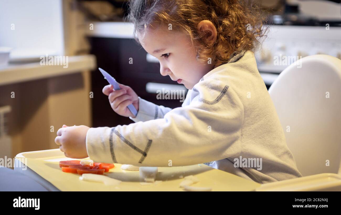 Kind schneidet Karotten und Kartoffeln in der Küche, hübsches kleines Mädchen kocht mit Spielzeugmesser, niedliches zweijähriges Baby spielt mit Essen zu Hause, glücklich unabhängig k Stockfoto