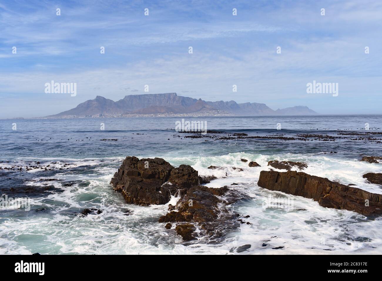 Tafelberg, Lions Head & Devils Peak von Robben Island aus gesehen, gegenüber der Table Bay. Der Ozean stürzt gegen die felsige Uferlinie. Stockfoto
