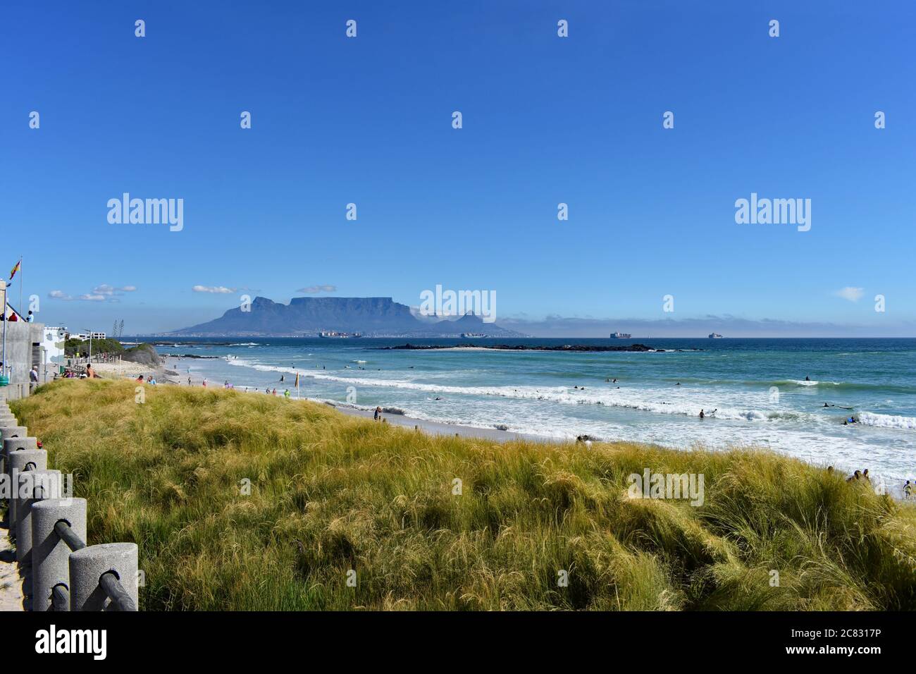 Tafelberg, Devils Peak & Lions Head vom Bloubergstrand Beach aus gesehen von der anderen Seite der Table Bay. Grünes Gras kontrastiert mit dem Blau des Himmels und des Ozeans Stockfoto