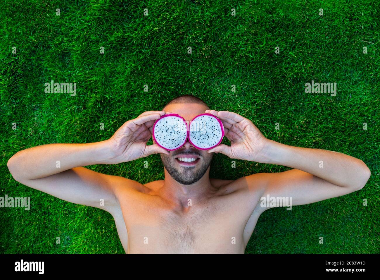 Lächelnder junger Mann, der auf einem grünen Gras liegt, mit Drachenfrucht statt mit Augen Stockfoto