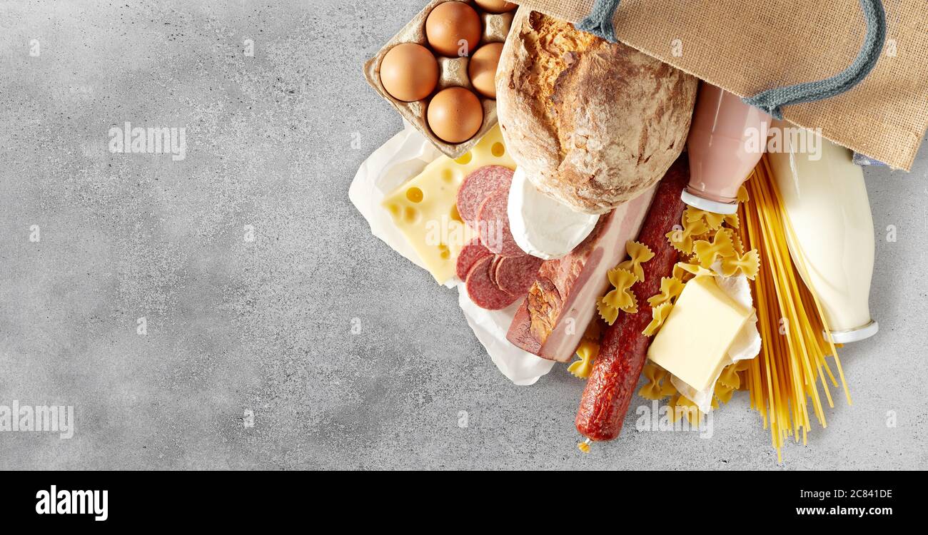 Verschiedene Milchprodukte mit Eiern, Pasta und Salami, die aus einem wiederverwendbaren Einkaufstasche auf einen strukturierten grauen Hintergrund mit Copyspace in einem Panorama verschüttet werden Stockfoto