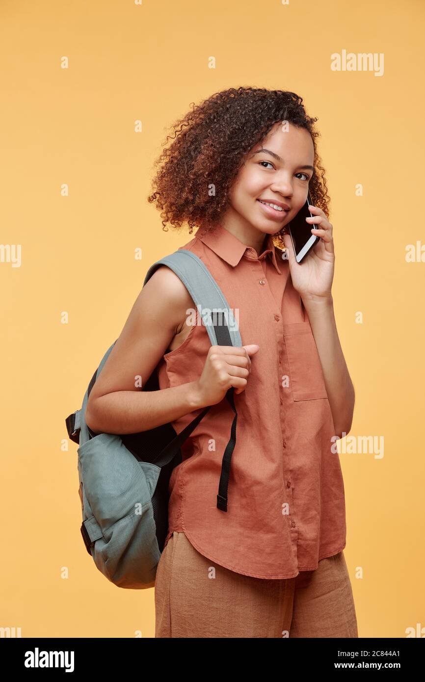 Fröhlich attraktives afro-amerikanisches Studentenmädchen in lässigem Hemd mit Tasche und telefonischer Telefonanruf gegen gelben Hintergrund Stockfoto